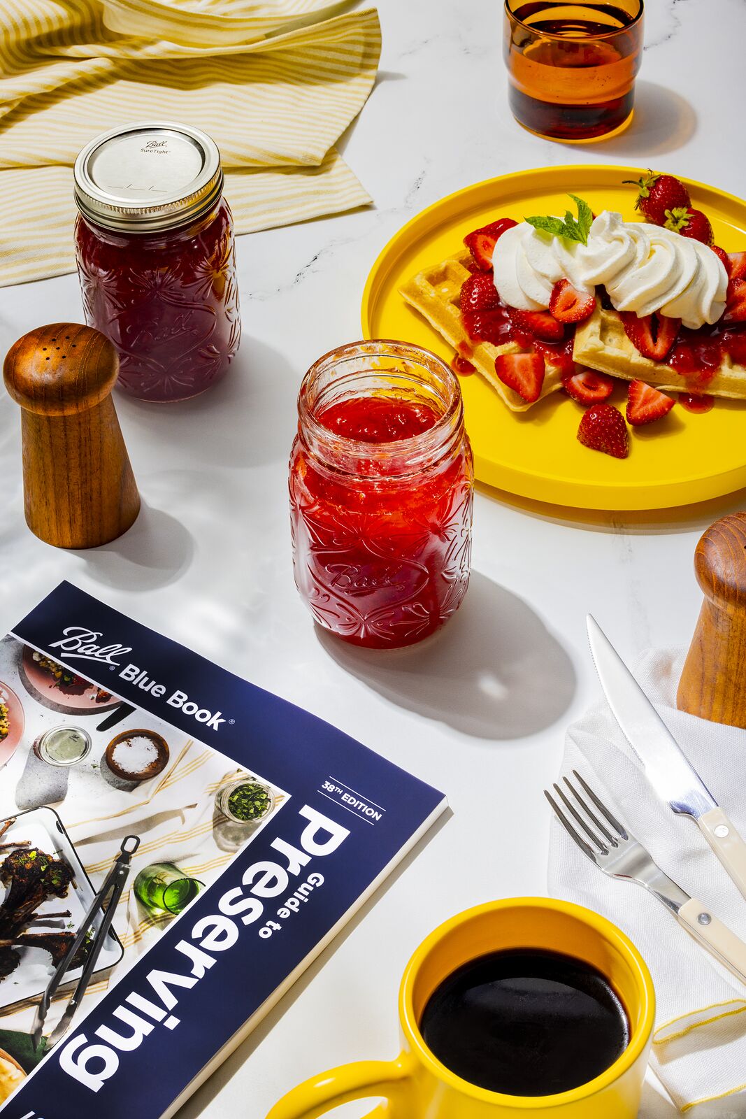 Breakfast spread with the Ball Blue Book on the table beside a jar of red jam