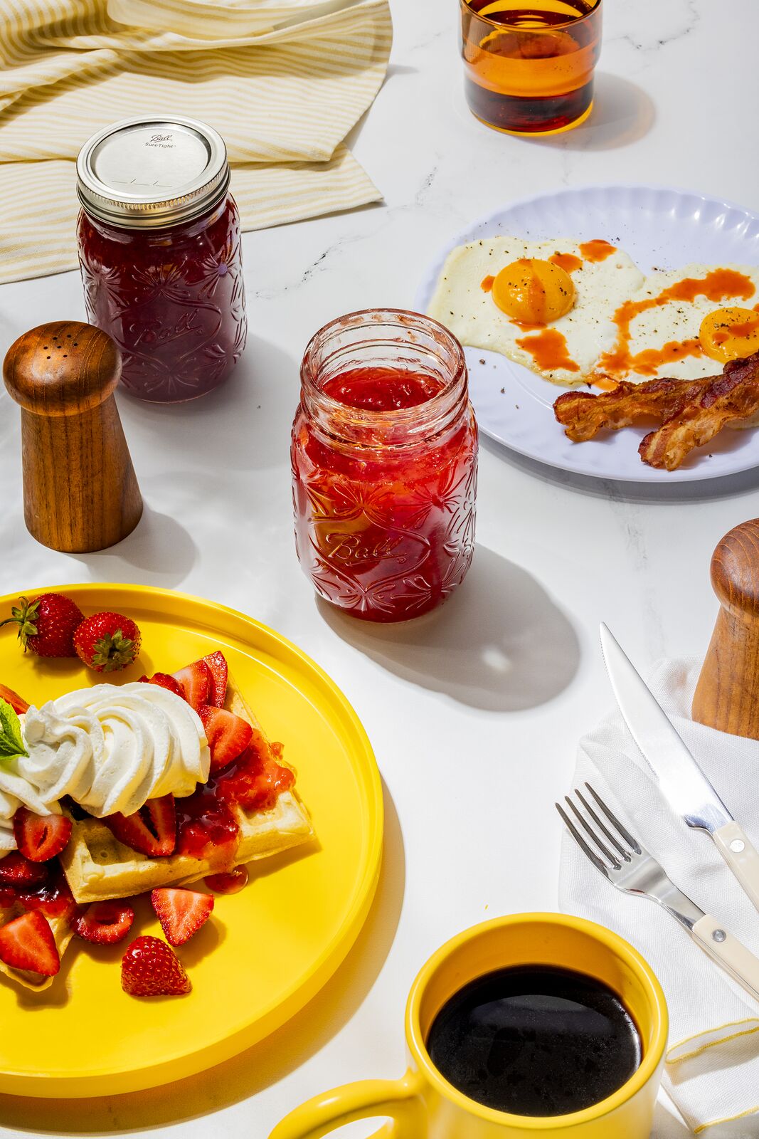 Breakfast table with a strawberry and cream waffle, bacon, coffee and a Ball jar of red jam
