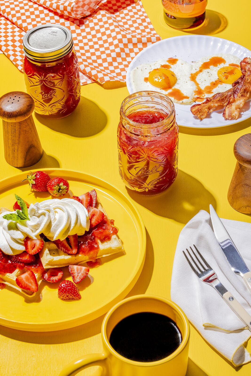 Breakfast on a yellow tabletop, waffle with strawberries and cream, bacon, and a jar of red jam