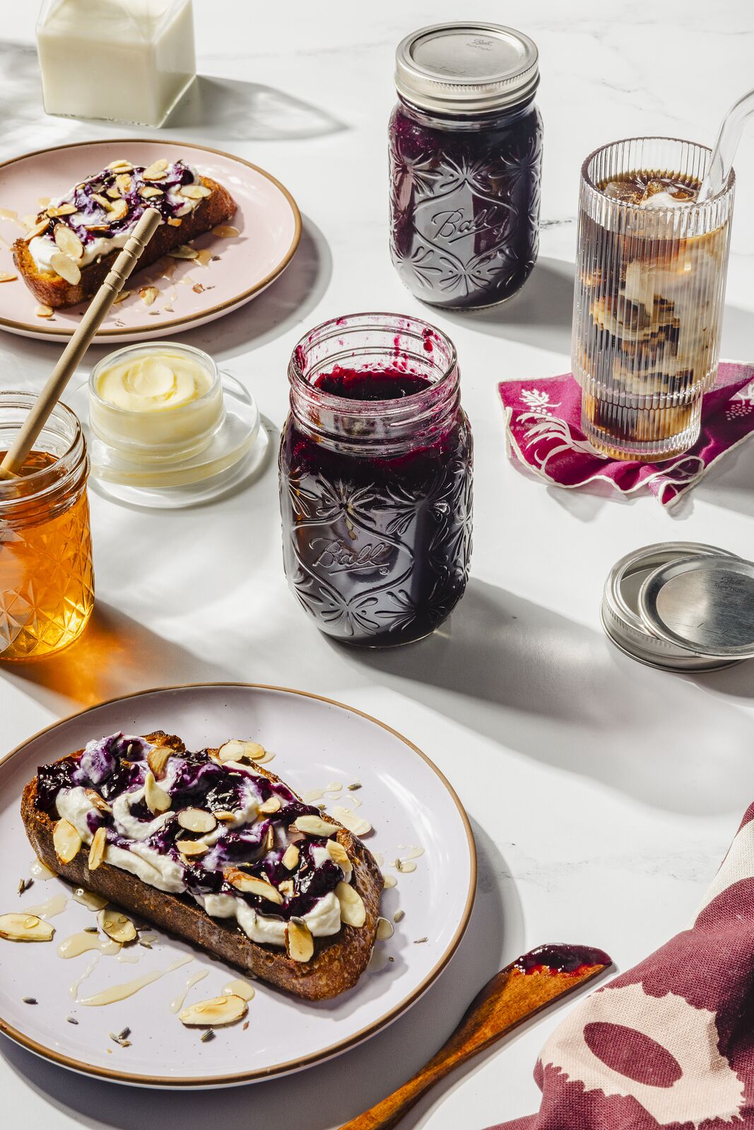 Brunch table with almond and jam toast, open jars of dark preserves, honey and a glass of milk