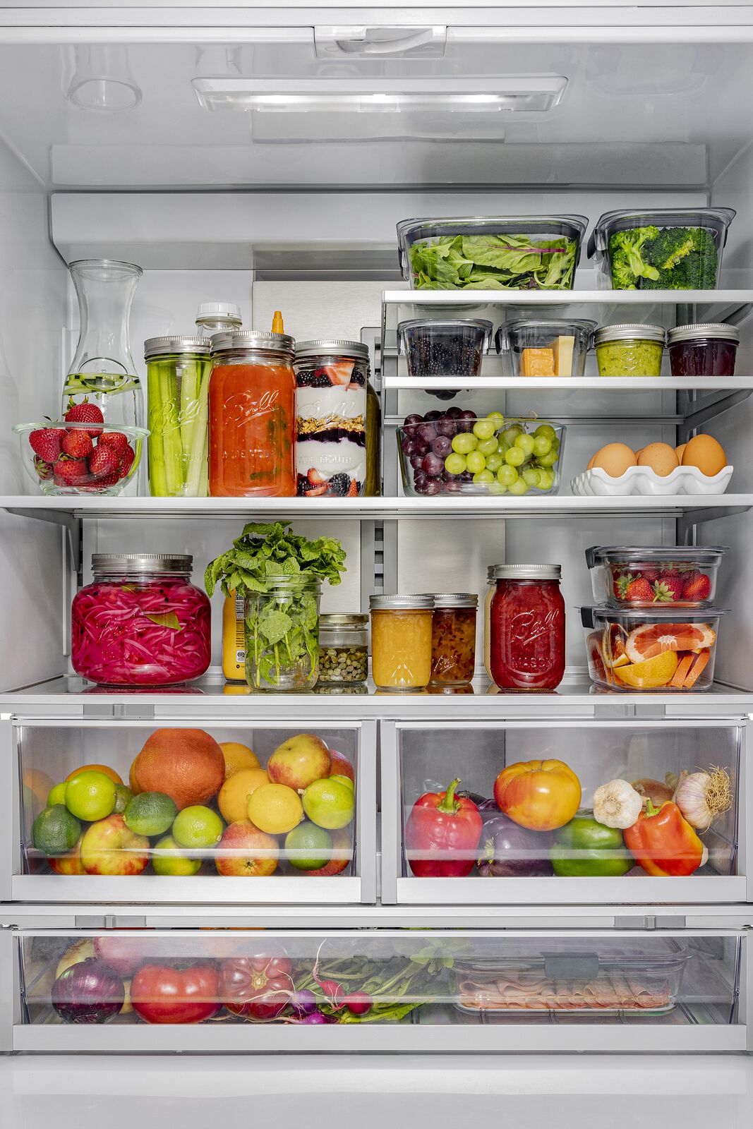 Refrigerator stocked with Ball jars of preserves and prepared food alongside fresh produce