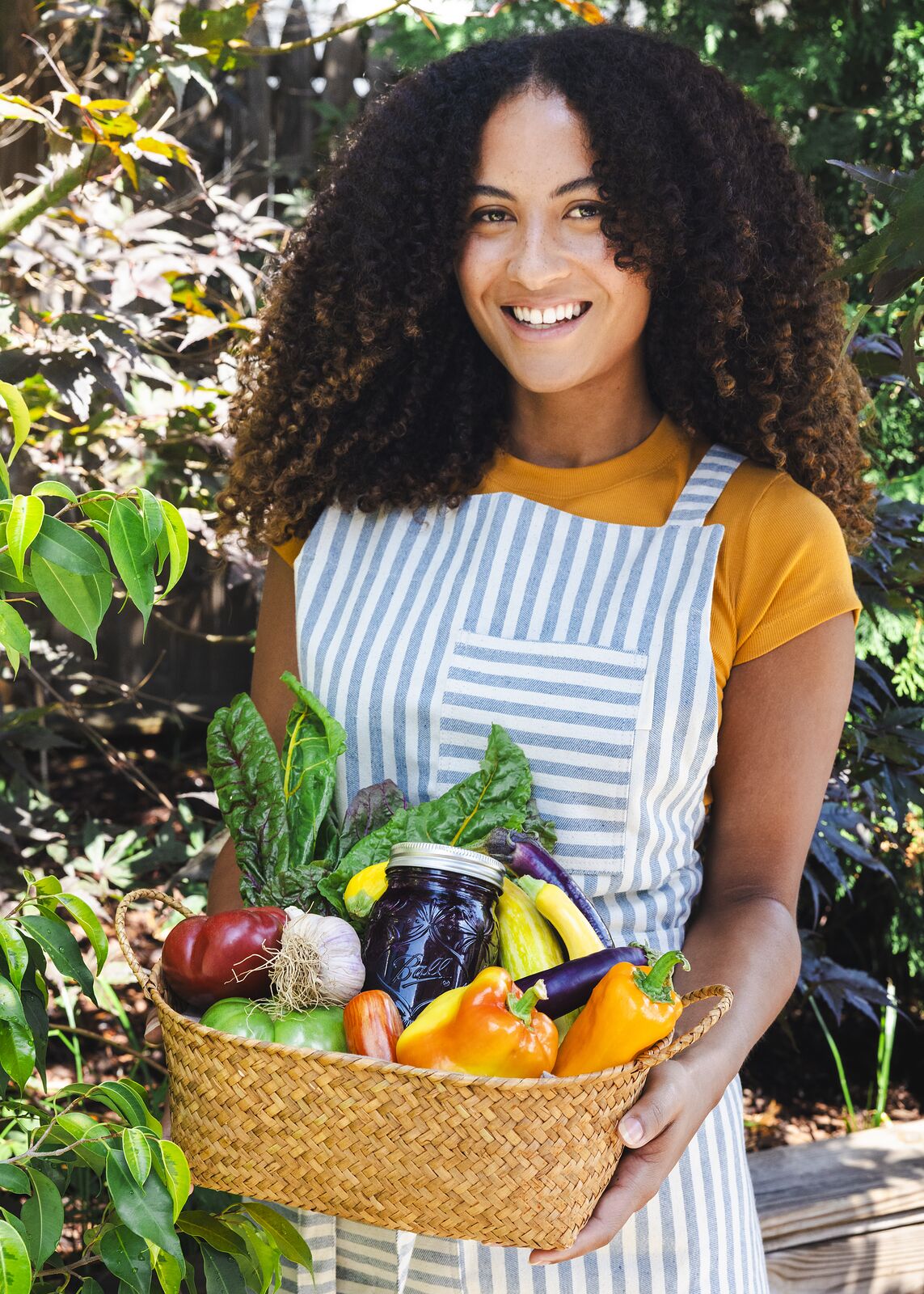 Gardener in a striped apron smiling with a basket of peppers, eggplant, chard and a sealed Ball jar of dark preserves