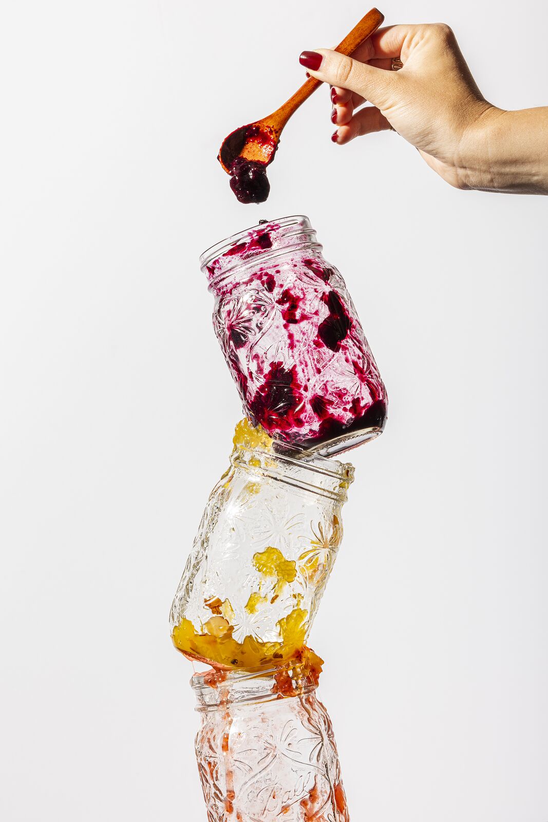 Spoonful of dark berry jam held above a stack of balanced Ball jars on a white background