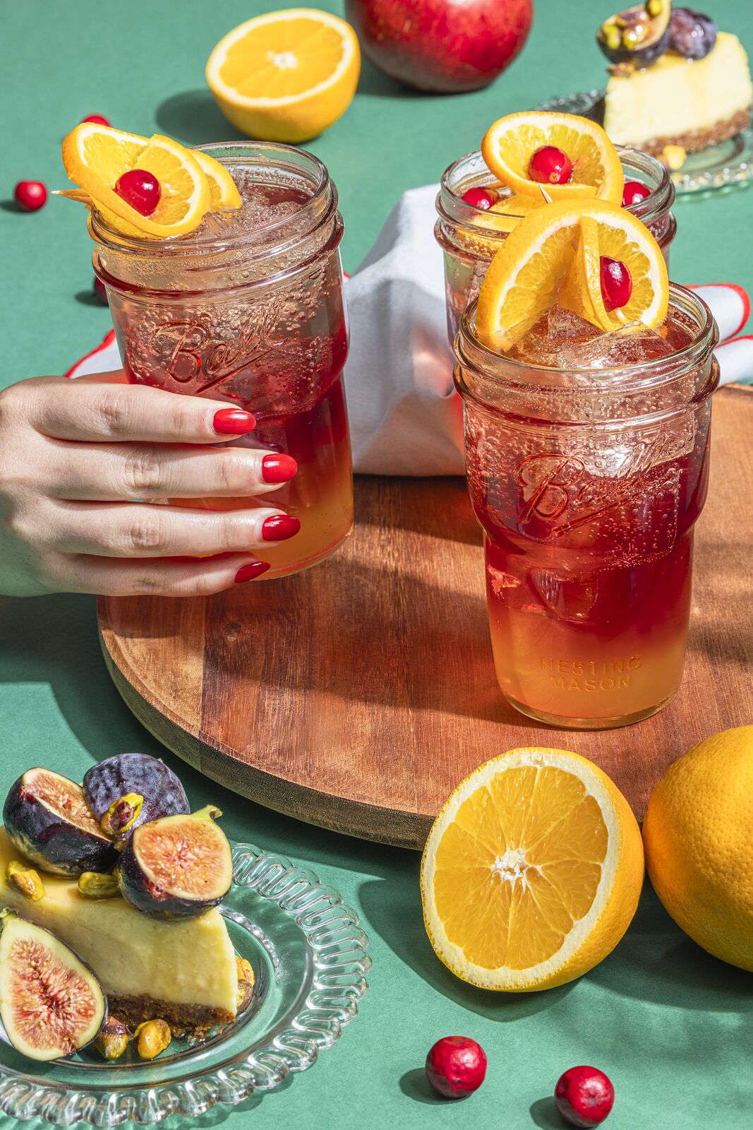 Hand resting on a Ball jar drink garnished with an orange twist, shot against a teal backdrop