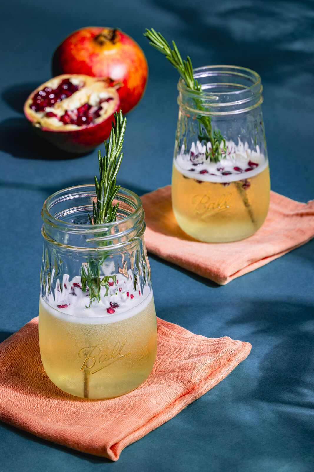 Two Ball jars of a foam-topped drink with rosemary and pomegranate on a deep blue set