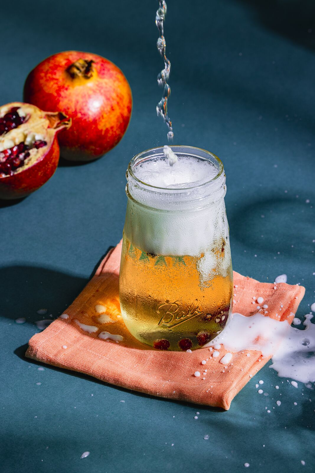 Drink poured into a Ball jar to a foaming head, pomegranate alongside on blue