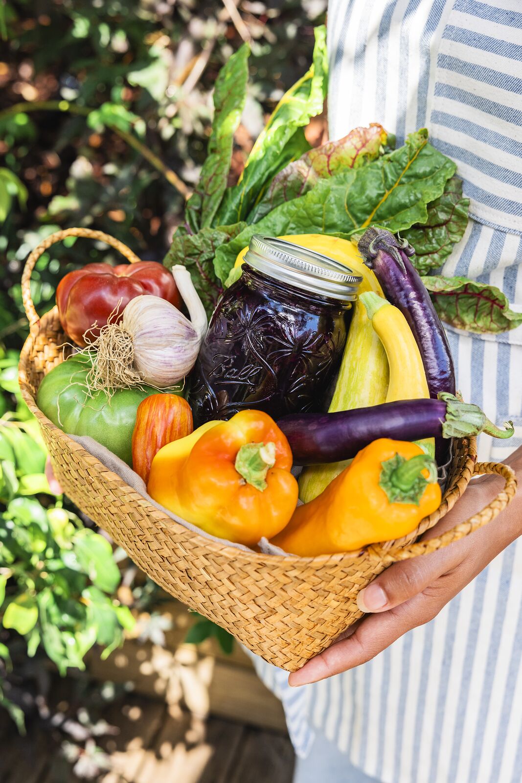 Woven basket of peppers, squash, eggplant and chard packed around a Ball jar of dark preserves