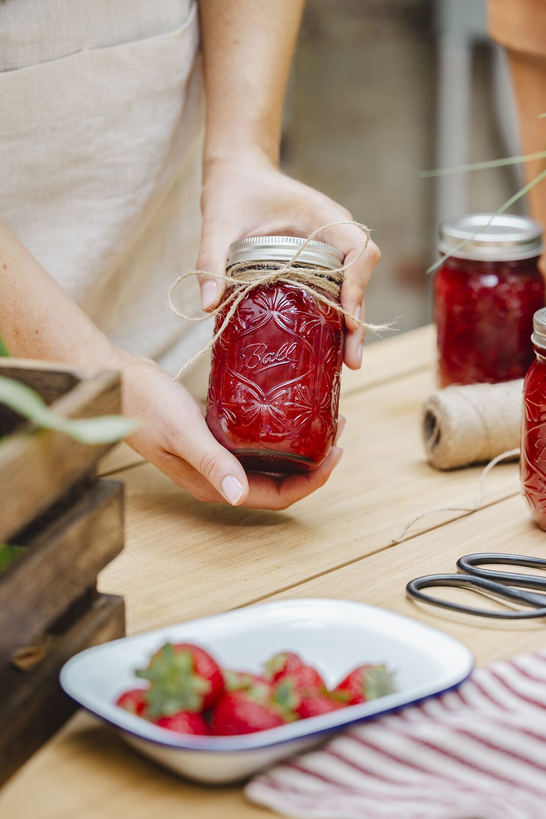 Hands tying twine around a sealed jar of red jam, fresh strawberries on an enamel plate