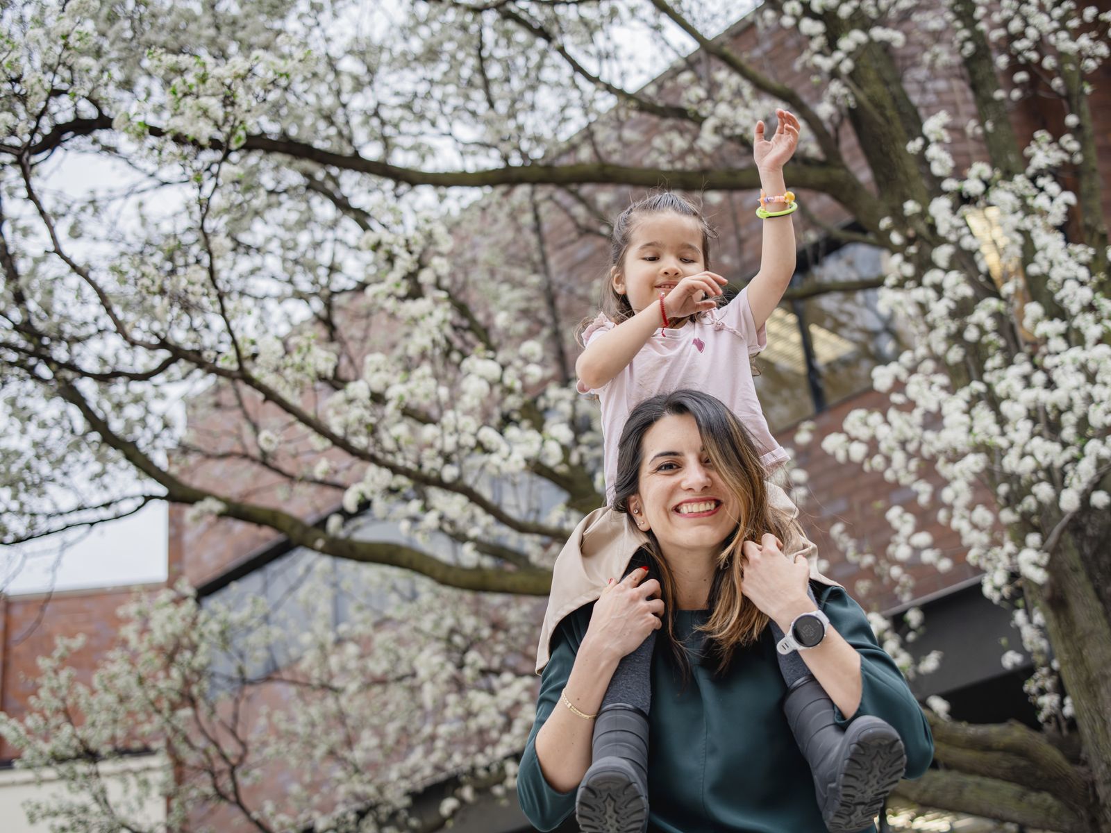 A woman in a dark green top smiles broadly while carrying a young girl on her shoulders along a city street. The girl, in a pale pink shirt with a heart on it and a stack of neon bracelets, has one arm flung straight up. The branches of a pear tree in full white blossom fill the frame above and behind them.
