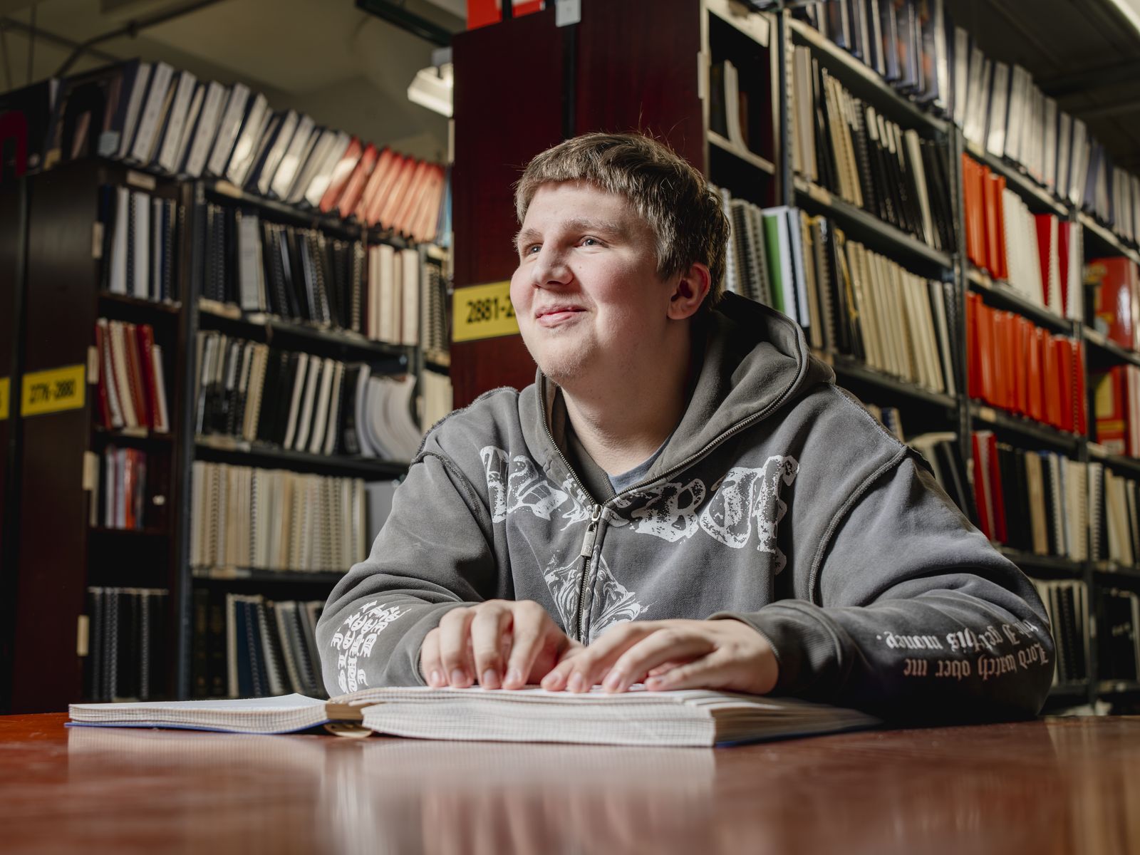 A teenage boy sits at a wooden reading table with both hands resting flat on an open braille volume, looking up and away from the page with a small smile. Behind him, floor to ceiling shelves hold hundreds of bound braille books in white, black and orange spines, with yellow catalog numbers on the shelf edges.