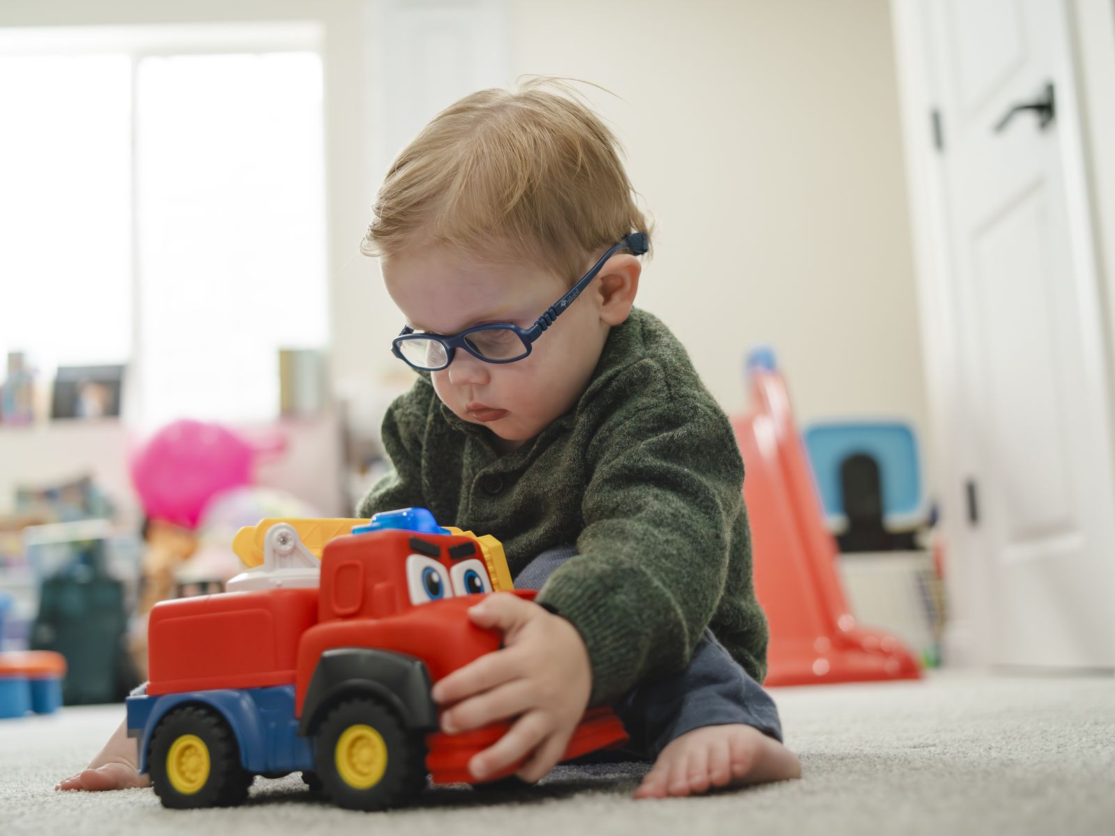 A blond toddler in navy blue glasses kneels on a bedroom carpet with both hands on a red and blue toy fire truck, head tipped down and fully absorbed in it. A slide and toy bins sit out of focus behind him in the afternoon light.
