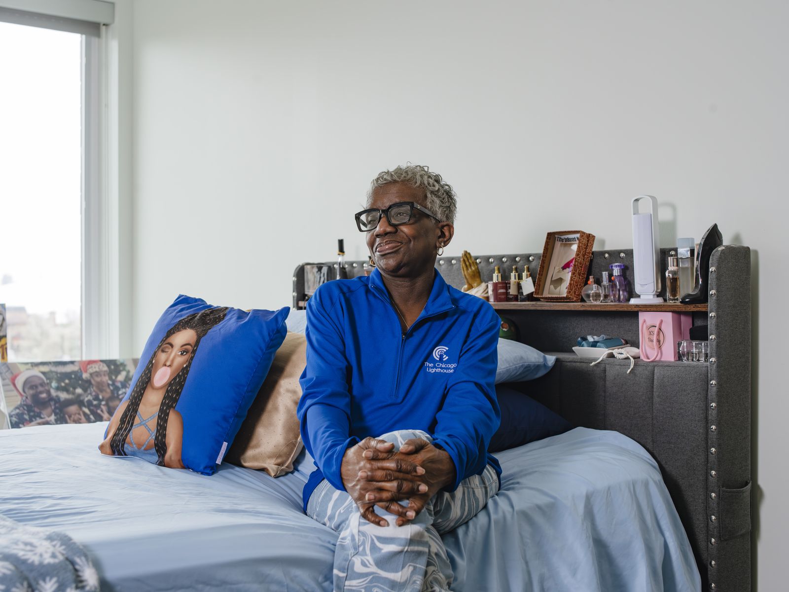 A woman with close cropped gray hair and heavy black glasses sits on the end of her made bed in a bright apartment, hands folded on her knee, wearing a royal blue Chicago Lighthouse quarter zip. Perfume bottles and framed photographs line the shelf behind the headboard and a window shows the city beyond.