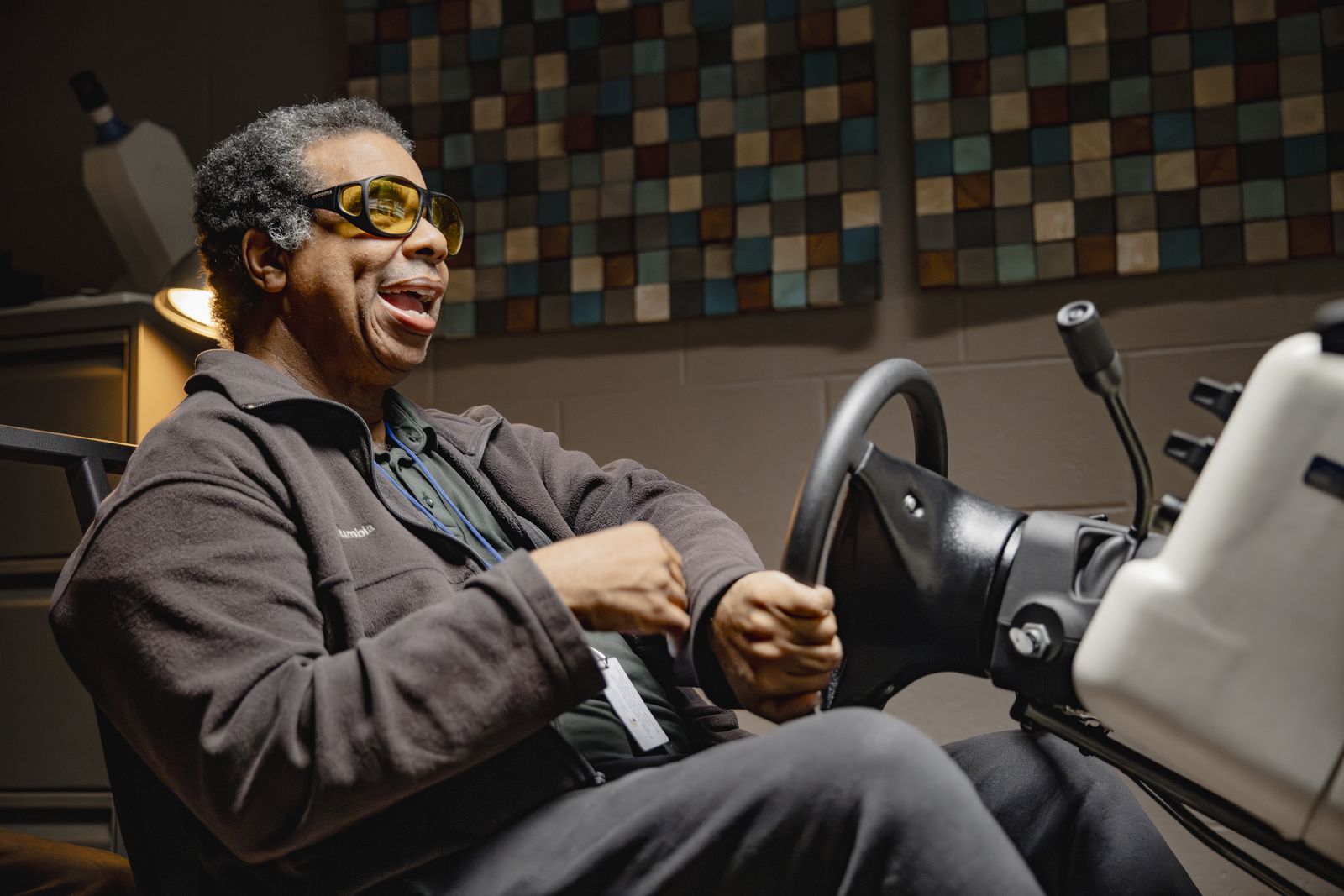 A man with gray hair, wearing amber tinted filter glasses, throws his head back laughing with both hands on the wheel of a driving simulator. The rig's screen and controls are lit beside him in a dim training room.